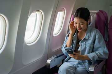 Asian traveler women surfing social media on smartphone and wearing bluetooth headphones to watching movie while sitting near windows during flight business travel in airplane with vacation lifestyle