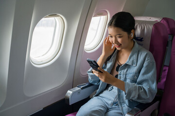 Asian traveler women surfing social media on smartphone and wearing bluetooth headphones to watching movie while sitting near windows during flight business travel in airplane with vacation lifestyle