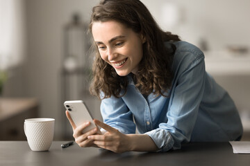 Pretty young woman holds modern gadget at home, leaned at workplace desk distracted from study or work chatting with friend, scroll news feed, spend time using social media platform mobile application