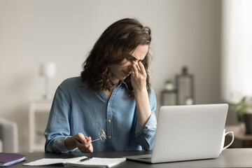 Tired woman sit at desk, takes off glasses to relieve eye strain after long laptop usage, suffers...