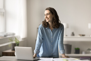 Cheerful young female standing at workplace desk, staring into distance, thinking feels happy with...