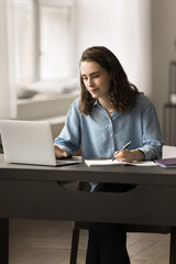 Focused school girl sits at table studying with laptop, preparing for college test or high-school...