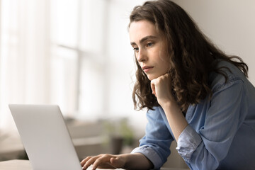 Pensive young woman stands at desk looking at laptop screen, learn new application, make...