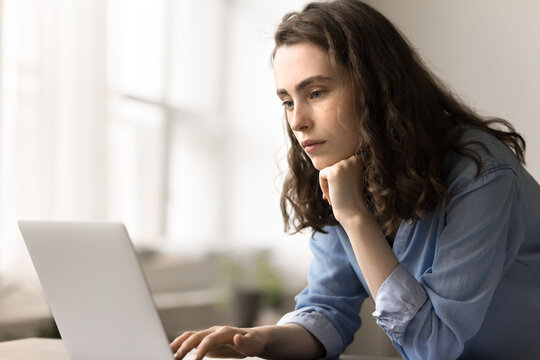 Pensive young woman stands at desk looking at laptop screen, learn new application, make assignment, search solution, consider idea, work on research project, looks focused while read on-line document