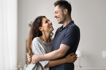 Beautiful millennial loving couple hugging, standing together in living room look at each other with love and warmth feel happy, enjoy their strong marriage, mutual understanding, harmonic relations