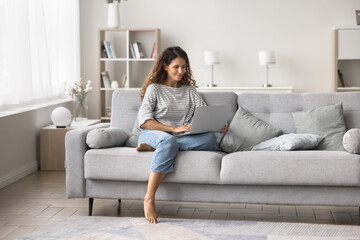 Young Hispanic woman relaxing on sofa in living room with laptop, spend weekend on internet, enjoy on-line communication, chatting in social media networks, share news, writes message, ordering goods