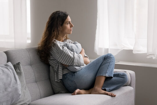 Serious pensive young woman thinking on problem, hugs cushion seated on couch at home, deep on sad thoughts, staring out window, looks depressed, frustrated, misses her beloved, feels lonely. Solitude