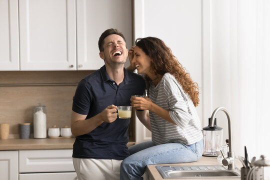 Happy Hispanic loving wife and husband spend time together in modern kitchen, laughing, drinking juice, talking, share funny life stories. Carefree spouses enjoy leisure at home. Family pastime, love