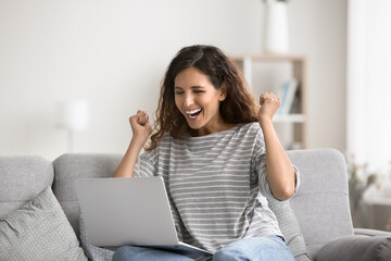 Happy woman sits on couch at home with laptop, check email, get great fantastic news, screams with joy, make gesture of winner, celebrate success, relish moment of lottery win, receive on-line prize