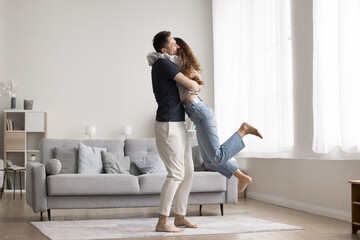 Couple celebrate relocation to new modern apartment, loving husband lifts his overjoyed wife,...