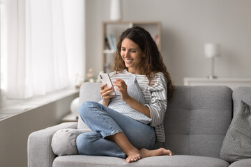 Smiling Latina woman resting on cozy sofa with smartphone, making call, check personal organizer using mobile application, enjoy remote communication with friend, ordering delivery services at home