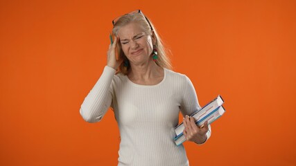 Portrait of frustrated, tired blonde teacher woman 40s 50s years old standing hold books massaging temples isolated on bright orange color background studio