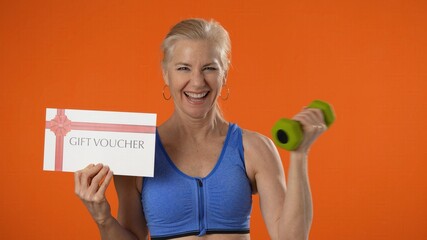 Smiling mature woman working out exercise with dumbbell holding a gift certificate voucher on orange background with copy space studio.