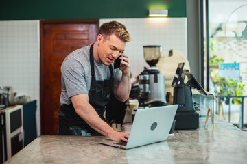 Young men barista wear apron talking telephone and using laptop computer at coffee shop. male staff standing at counter receive customer orders in cafe. business coffee store concept.