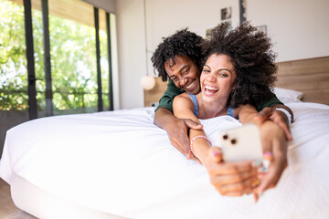 A young couple of African descent is lying on a bed in a modern, sunlit bedroom, smiling and...