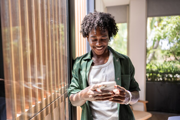 Young cheerful man of African descent standing near a big sunlit window of a hotel room, looking at...