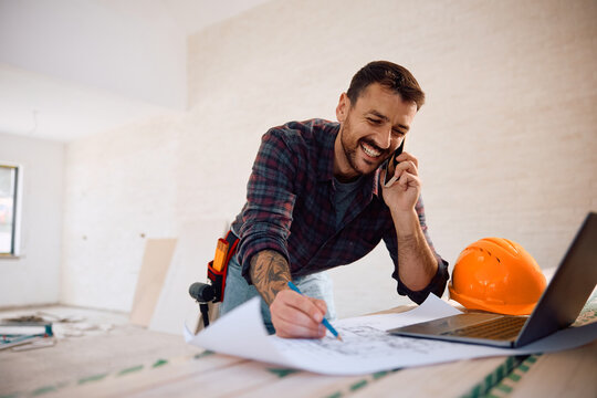 Happy worker talking on the phone while examining housing plans at construction site.