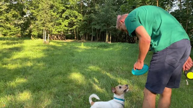 A young man throws a plastic frisbee for a dog. The animal trainer is teaching tricks to the dog at a public park.