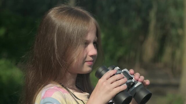 Kid girl with binoculars on green background close up. Happy child scout outdoors. Concept of summer camp, adventure, scouting and hiking tourism for children