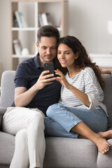 Fototapeta premium Hispanic spouses rest on cozy sofa looking at mobile phone screen, web surfing information, buy tickets, booking hotel, enjoy new modern application, communicate using wireless internet. Technology