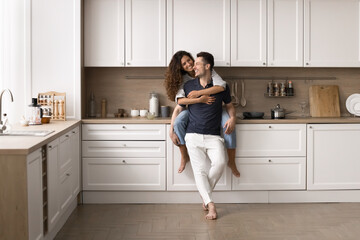 Happy beautiful wife sits on countertop, cuddling husband, smile, enjoy talk, spend time together in good furnished kitchen. New house, bank mortgage for young family, renovation, remodeling services