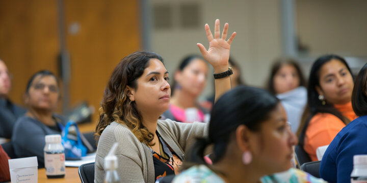 hispanic woman raises her hand in a classroom setting, asking a question to the lecturer or teacher.