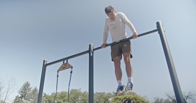 An athletic man performs pull-ups on parallel bars in an outdoor park setting with clear skies, showcasing his strength and commitment to fitness.