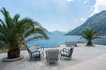 Serene waterfront patio featuring a set of cushioned chairs around a coffee table, with palm trees, overlooking a calm bay surrounded by mountains