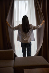 Obraz premium Slim black-hair woman standing against big window and opening curtains
