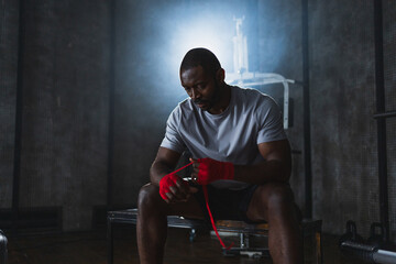 Fitness workout in gym. African American man fighter preparing for fight wrapping hands with red boxing wraps sports protective bandages in gym. Strong man ready for fight boxing sparring training