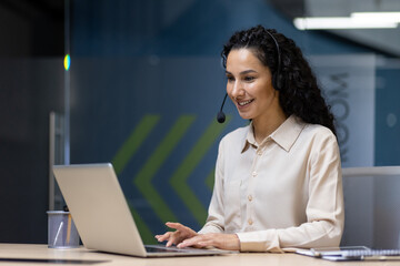 Smiling customer service representative engaging with clients via headset while using laptop. Modern office environment, professional attire, bright and welcoming atmosphere.