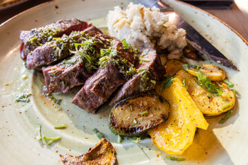 A plate of meat with a side of mashed potatoes and vegetables. The meat is cooked and seasoned perfectly, and the vegetables are roasted.
