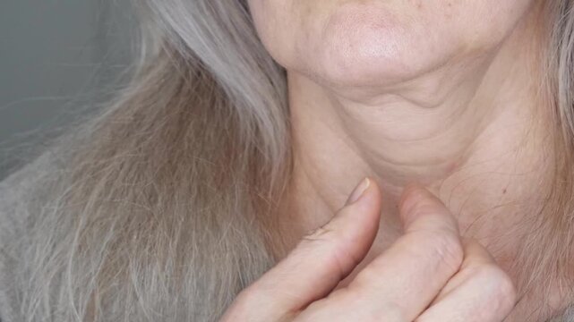 Close-up of a woman's neck with gray hair. The woman touches her loose and saggy skin on her chin and neck. The concept of cosmetology and plastic surgery. The concept of skin care in middle age.