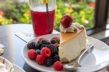 A slice of cheesecake with strawberries, blueberries and raspberries on top of a white plate. A glass of red juice is next to the plate