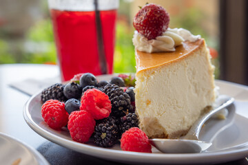 A slice of cheesecake with strawberries, blueberries and raspberries on top of a white plate. A glass of red juice is next to the plate