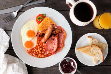 Full English breakfast with baked beans, bacon, eggs, sausage and hash browns. Overhead view table scene on a dark wood background.