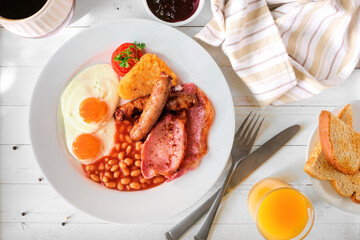 Full English breakfast with baked beans, bacon, eggs, sausage and hash browns. Top view table scene over a white wood background.