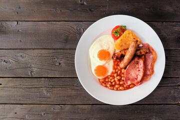 Full English breakfast with baked beans, bacon, eggs, sausage and hash browns. Top view over a rustic dark wood background.