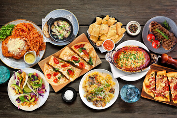 Table scene of various delicious foods. Top down view on a dark wood background. Pastas, flatbreads, tacos, steak and restaurant theme dishes.