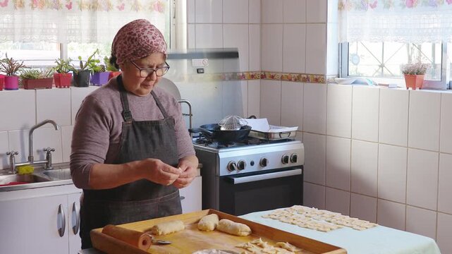 Senior latin woman kneading dough, cooking chilean fried sweet pastry calzones rotos in her domestic kitchen. 