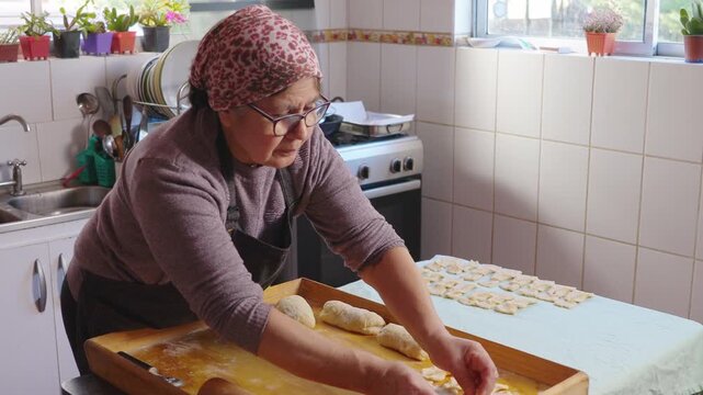 Senior latin woman kneading dough, cooking chilean fried sweet pastry calzones rotos in her domestic kitchen. 