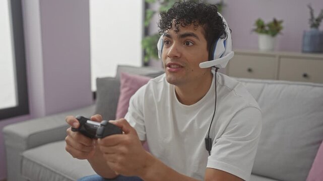 A focused young hispanic man with curly hair plays video games in a cozy living room, wearing headphones and holding a controller