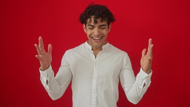Excited young hispanic man in shirt, standing isolated over red background, arms raised in victory, celebrating mad and crazy success as a winner