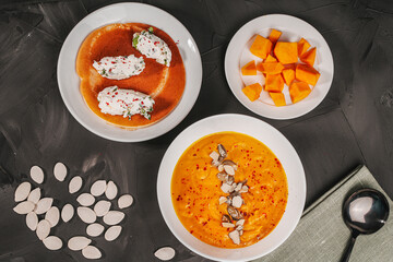 Traditional vegetable cream pumpkin and carrot soup in a white bowl, sliced pumpkin slices on a plate and pancake curd quenelles on a gray graphite background with pumpkin seeds