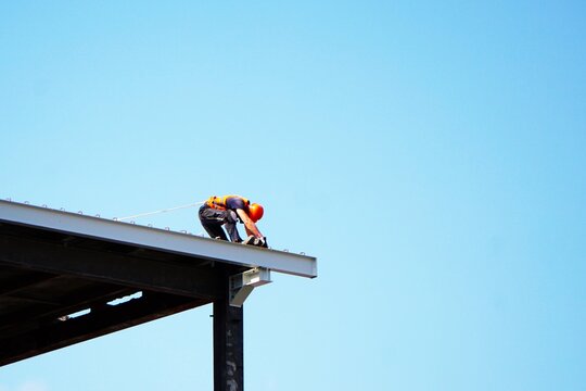 Mężczyzna ciężko pracuje na wysokości. Ciężka praca na budowie. Fizyczna praca na wysokości pod zabezpieczeniem. Budowlaniec w kasku i lina chroniąca pracownika przed upadkiem.