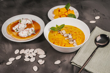 Traditional vegetable cream pumpkin and carrot soup in a white bowl, sliced pumpkin slices on a plate and pancake curd quenelles on a gray graphite background with pumpkin seeds