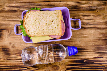 Bottle of water and lunch box with sandwiches on a wooden table. Top view