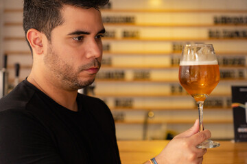 Young latin tasting craft beer in a pub, observing the color and hops in the lager.