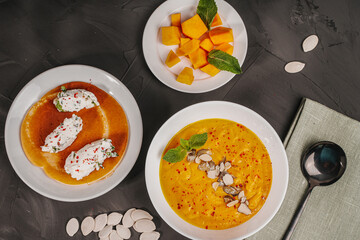 Traditional vegetable cream pumpkin and carrot soup in a white bowl, sliced pumpkin slices on a plate and pancake curd quenelles on a gray graphite background with pumpkin seeds