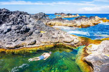 Unidentified man snorkelling in natural swimming pools on rocky coast in Mosteiros town, Sao Miguel...
